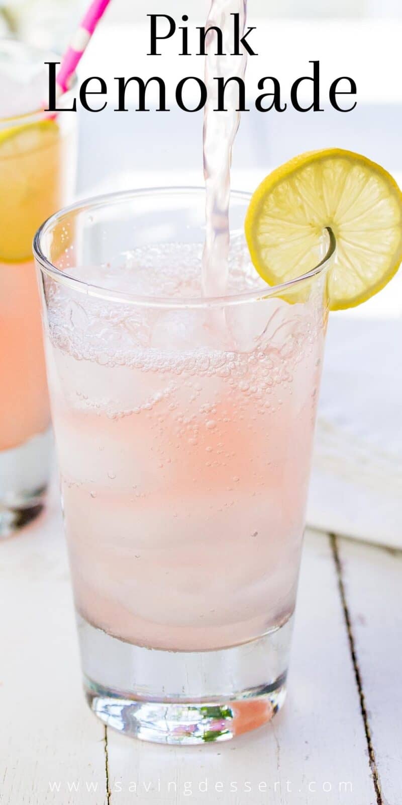 A tall glass being filled with homemade pink lemonade garnished with a slice of lemon