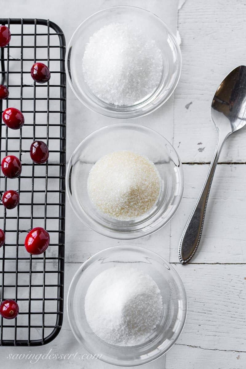 A rack of sticky cranberries next to three bowl of different kinds of granulated sugar