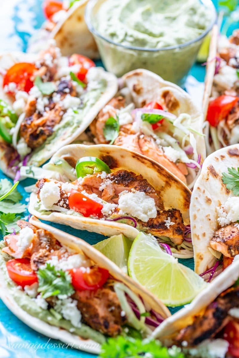 Grilled salmon tacos loaded with crunchy spicy slaw, a flavorful avocado crema and cheese garnished with lime wedges and cilantro