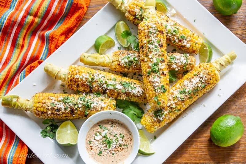 An overhead view of a platter of grilled corn with lime wedges and a dipping sauce