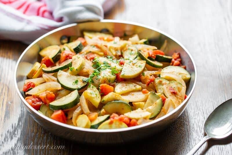 A side view of a skillet of summer squash, zucchini and onions with tomatoes