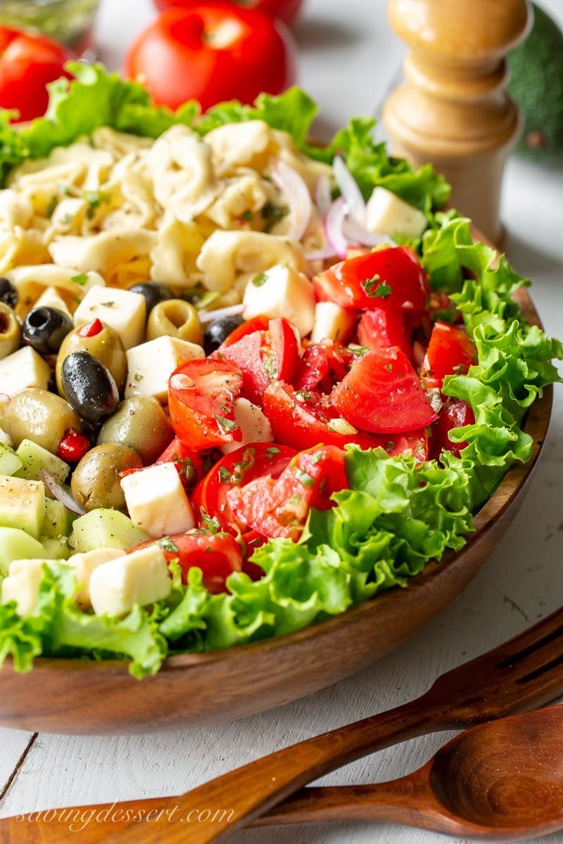 A large wooden bowl with Marinated Tomato & Tortellini Salad with cucumbers, onion, olives, avocado, cheese and fresh garden herbs. 