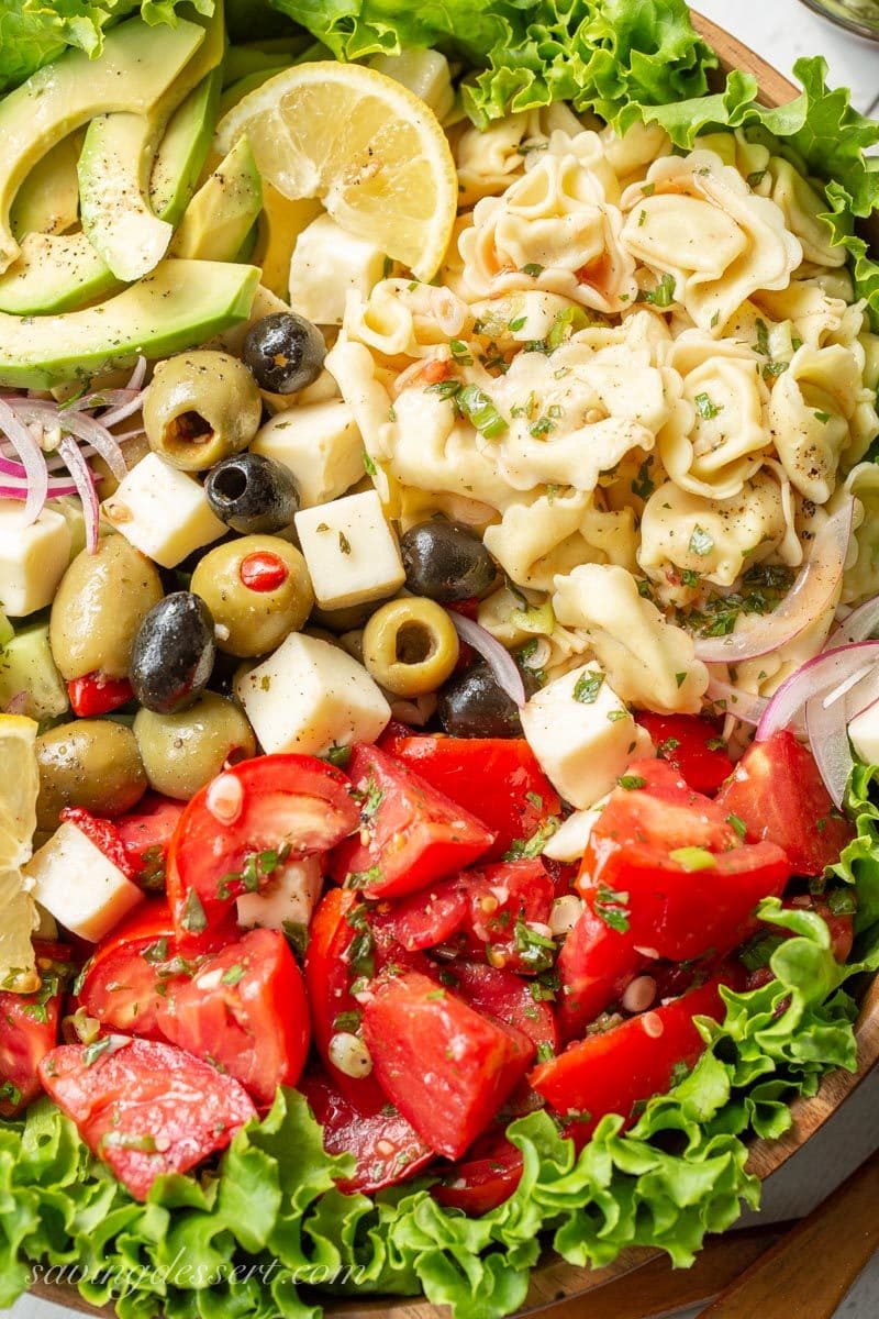 A large wooden bowl with Marinated Tomato & Tortellini Salad with cucumbers, onion, olives, avocado, cheese and fresh garden herbs. 