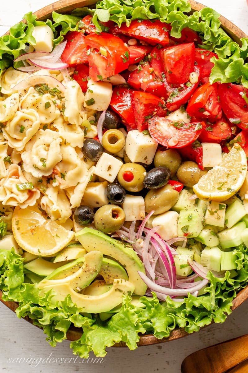 A large wooden bowl with Marinated Tomato & Tortellini Salad with cucumbers, onion, olives, avocado, cheese and fresh garden herbs. 