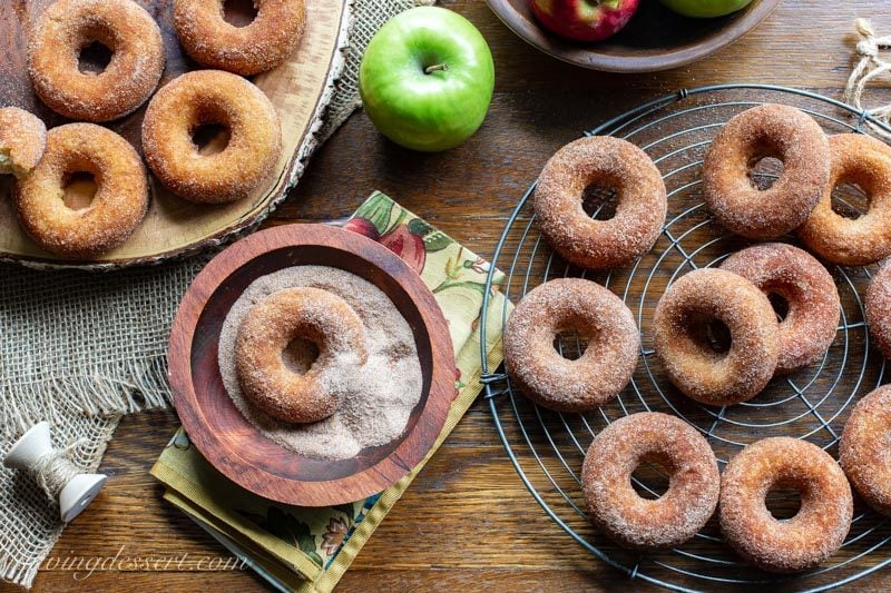 Baked apple cider doughnuts on a rack, board and one in a bowl tossed with a cinnamon sugar mixture