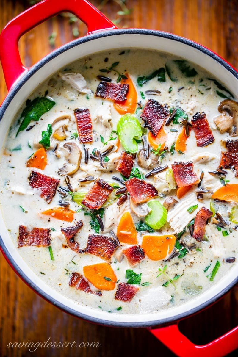 Red Dutch oven filled with creamy chicken and wild rice soup with carrots, celery, spinach and bacon on top