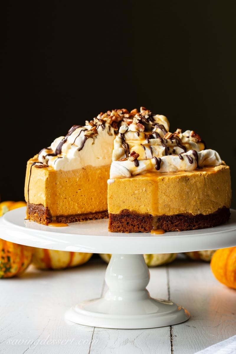 A sliced pumpkin cheesecake on a cake stand topped with whipped cream, chocolate and caramel