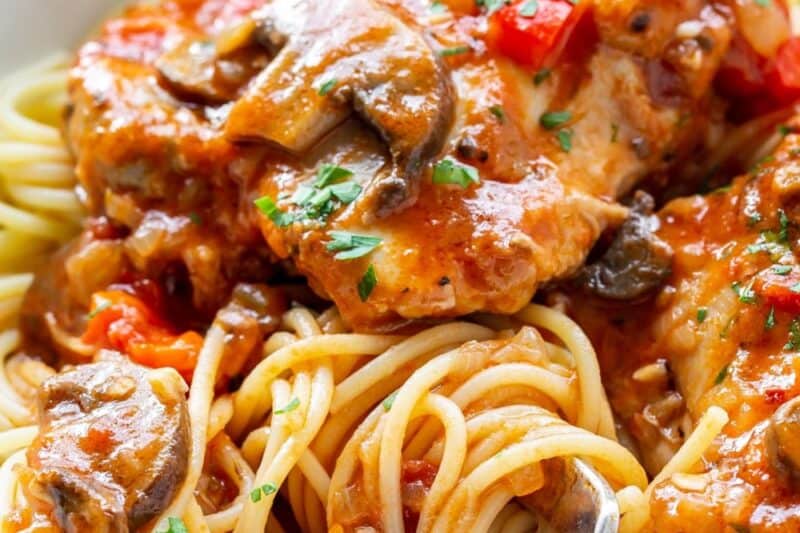 A side view of a bowl of spaghetti topped with chicken cacciatore with mushrooms and fresh parsley.