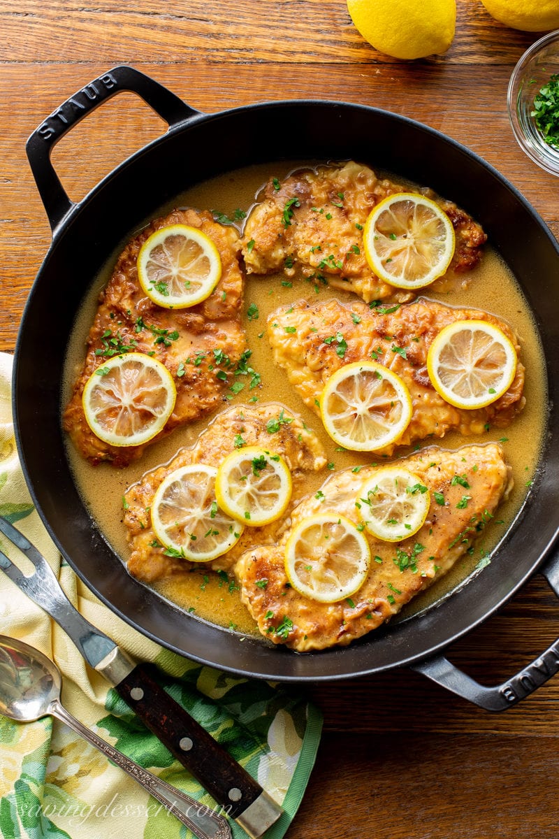 Classic Chicken Francese - A skillet with tender, breaded chicken cutlets in a bright lemon sauce garnished with parsley and sliced lemons. 