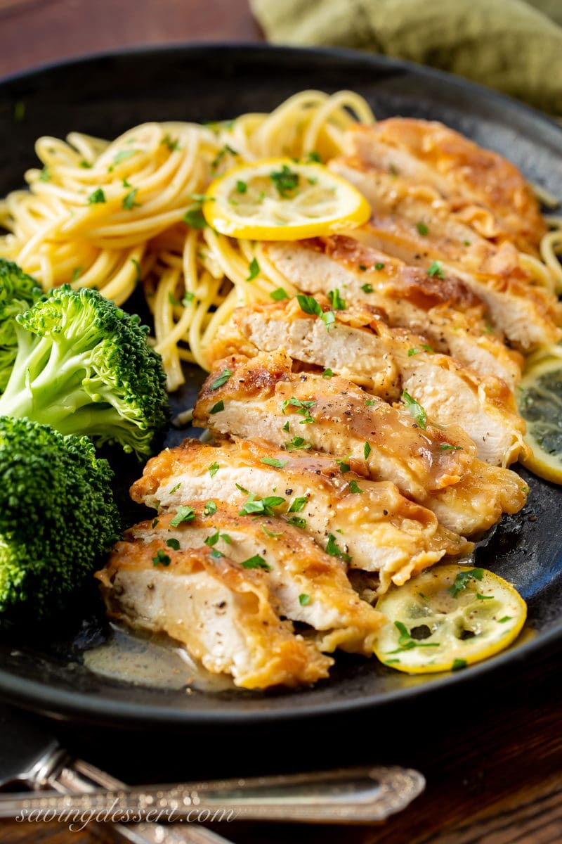 A dinner plate with steamed broccoli, spaghetti and sliced Chicken Francese garnished with sliced lemons, a buttery lemon sauce and fresh parsley