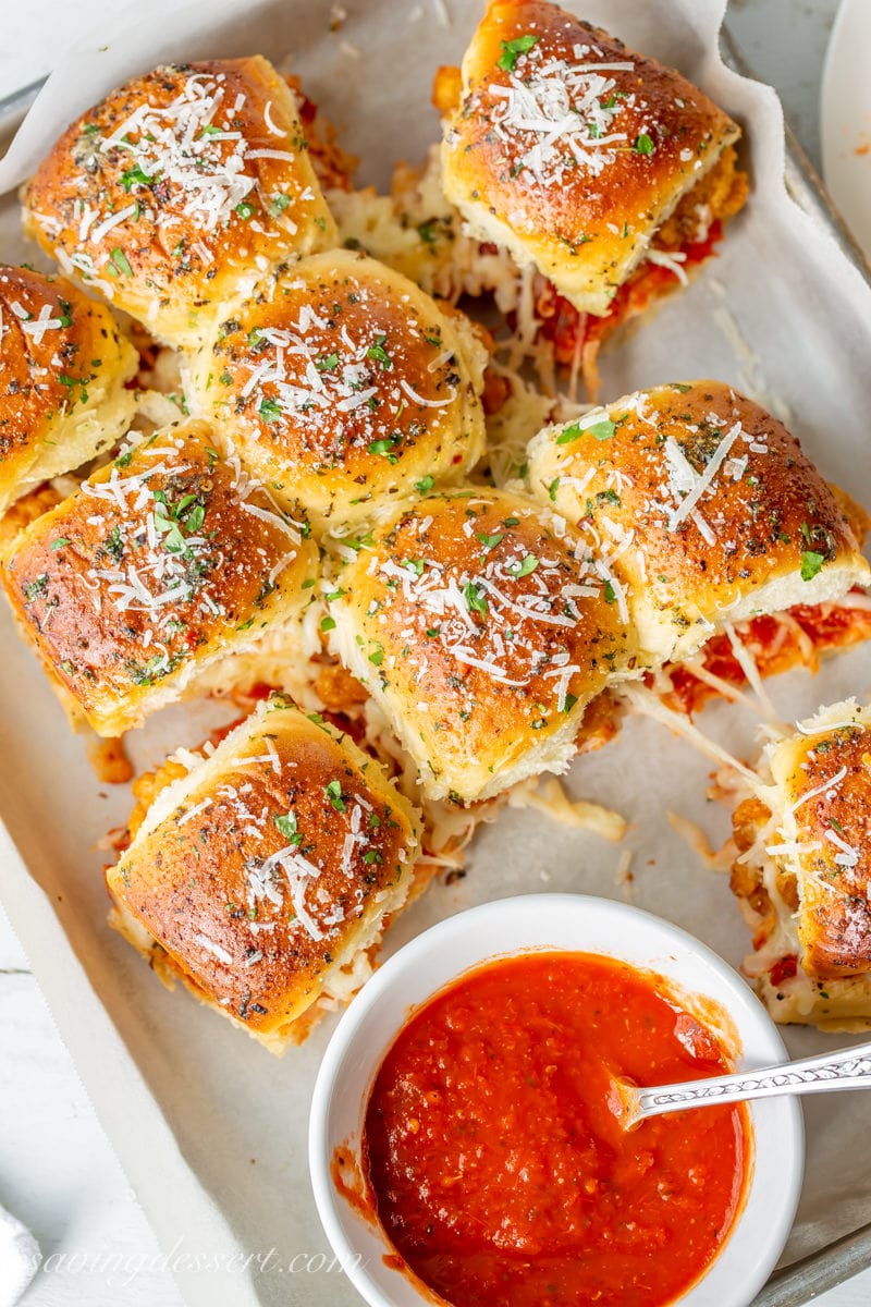 Overhead view of a tray of Chicken Parmesan Sliders with extra pizza sauce on the side. The rolls are brushed with a garlic herb butter sauce then topped with fresh parsley and grated Parmesan