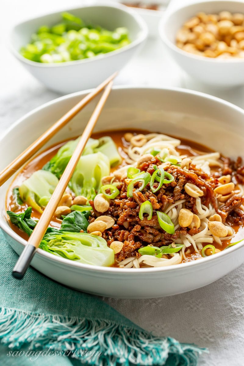 A big bowl of spicy Dan Dan Noodles with minced pork, bok choy and a rich peanut sauce garnished with peanuts and sliced green onions.