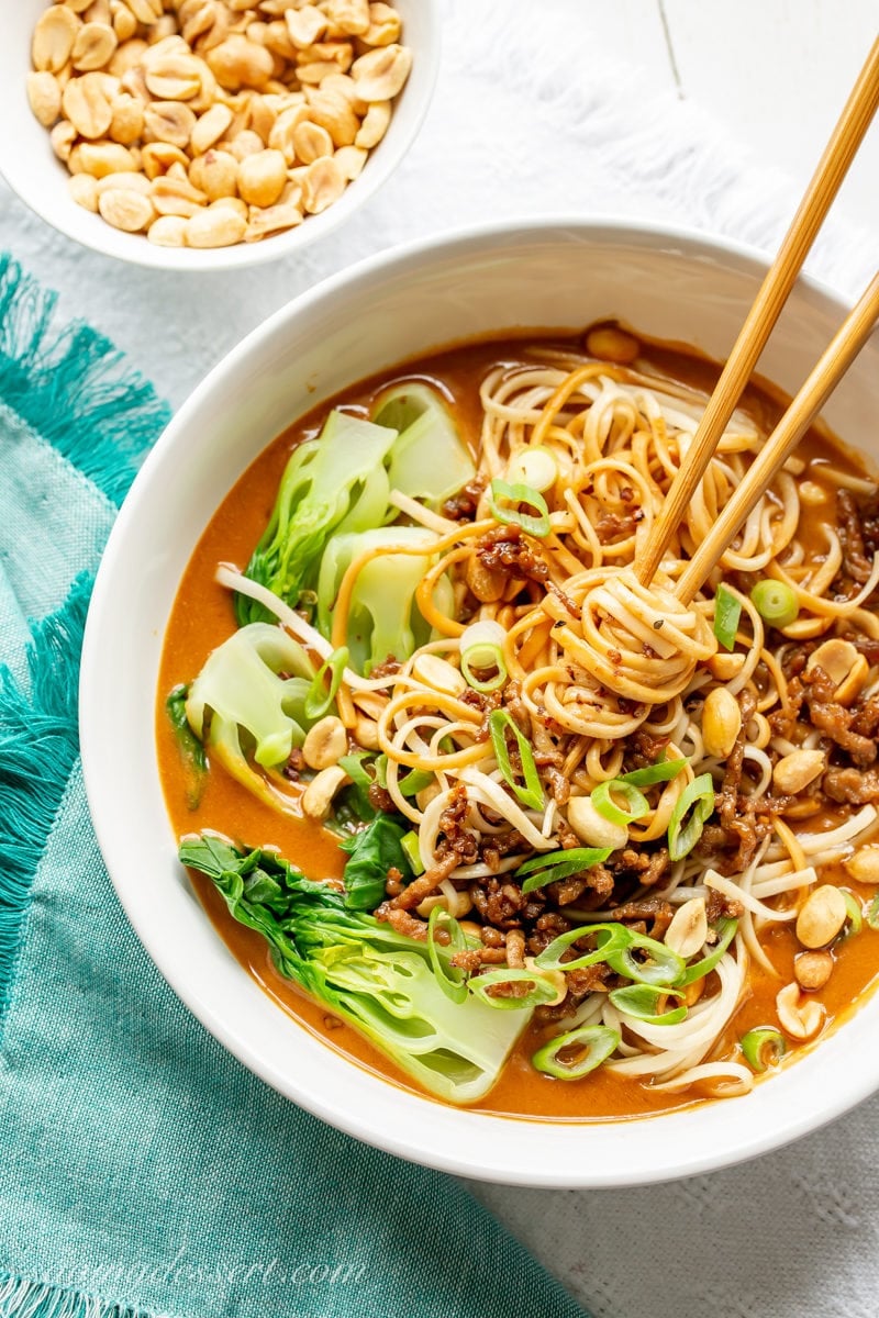 A bowl of spicy Dan Dan Noodles with chop sticks, bok choy, sliced green onions and peanuts