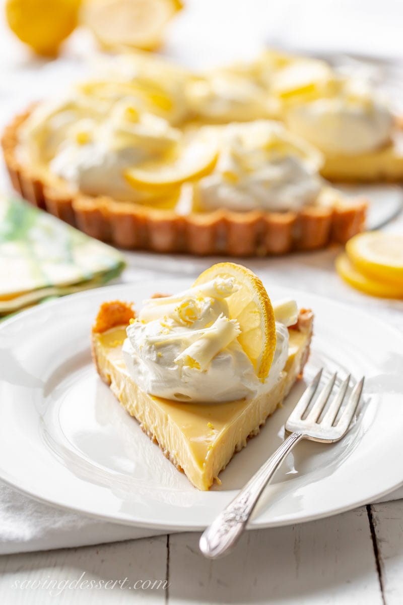 A slice of lemon pie with a mound of whipped cream, sliced lemon and white chocolate curls.