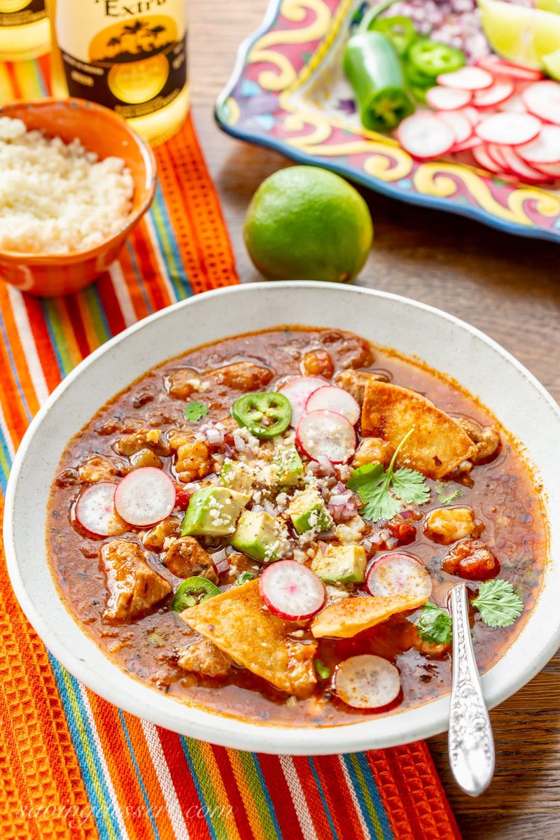 A bowl of pork posole with loads of garnishes like jalapeño slices, corn chips, sliced radishes and avocado