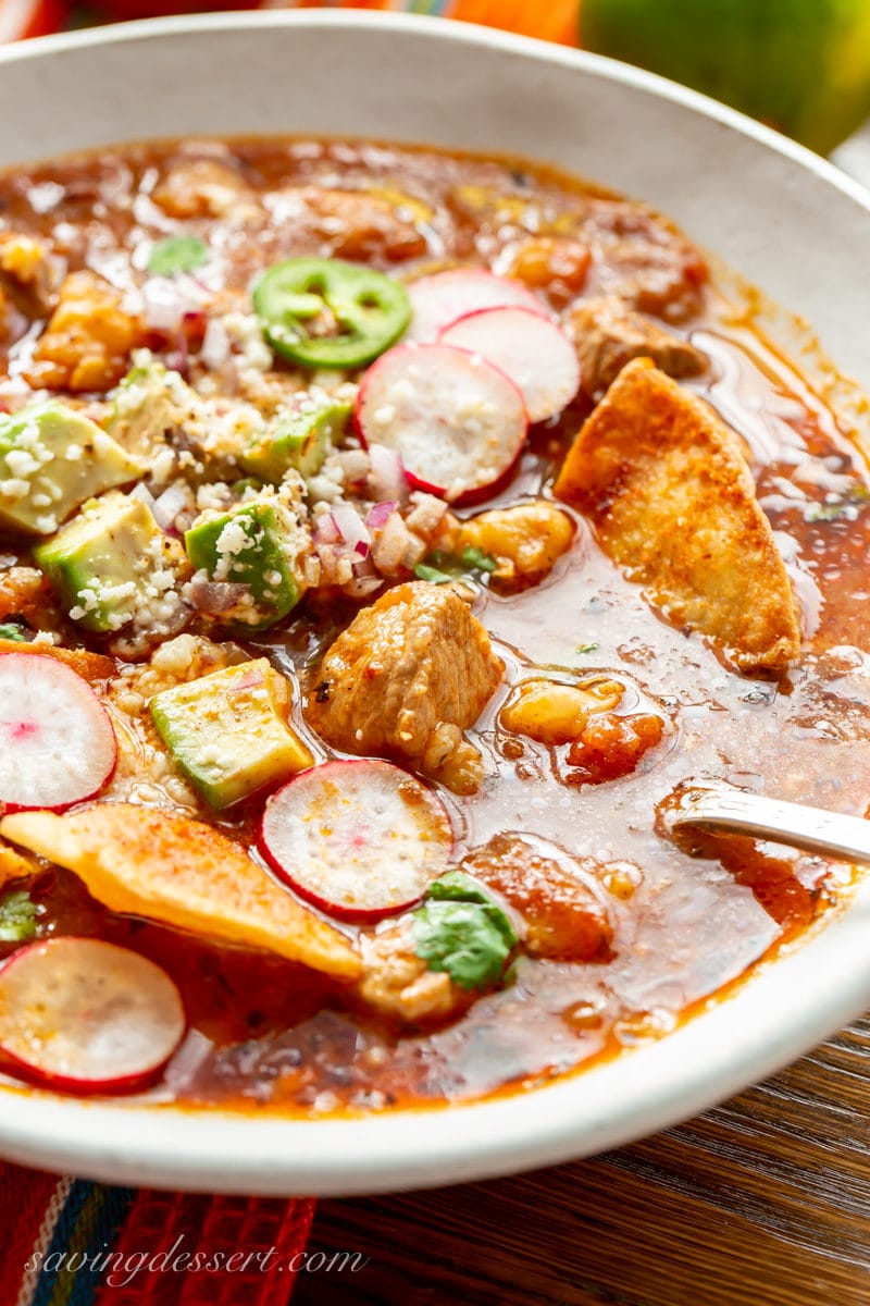 Close up of a bowl of pork posole with tender chunks of pork in a rich broth topped with sliced radishes, cheese, cilantro and avocado