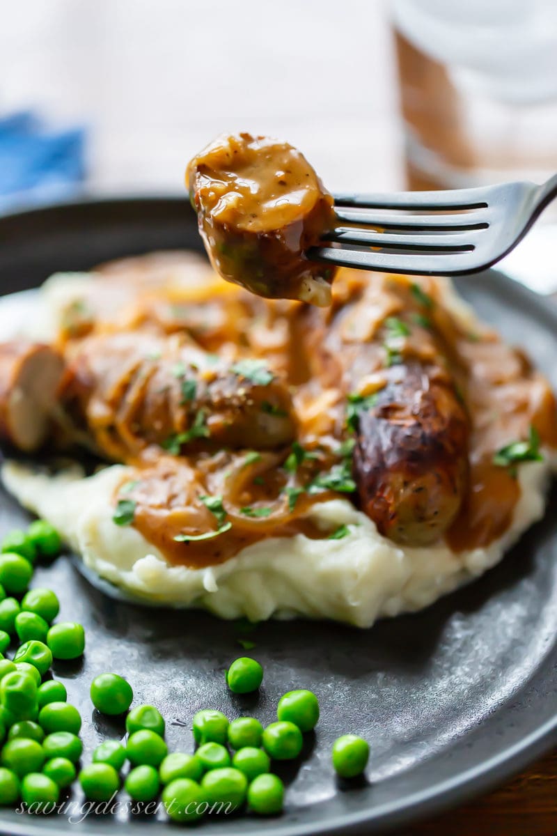 A fork with a slice of gravy covered sausage from a plate of classic Bangers and Mash served with peas
