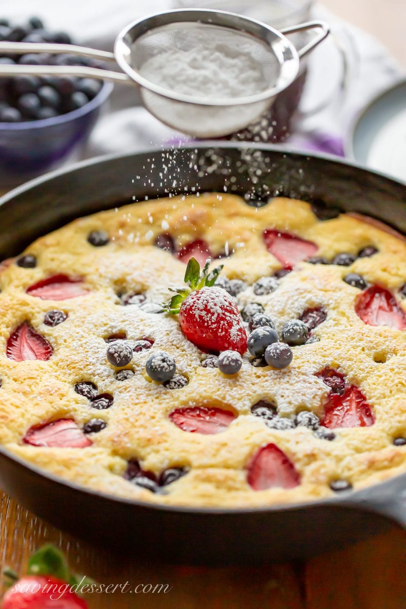 A cast iron skillet with a baked pancake with blueberries and strawberries being covered with sifted powdered sugar