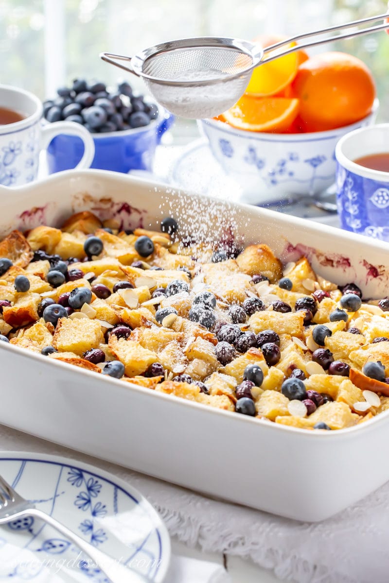 A baking dish with an overnight blueberry french toast casserole being dusted with powdered sugar.