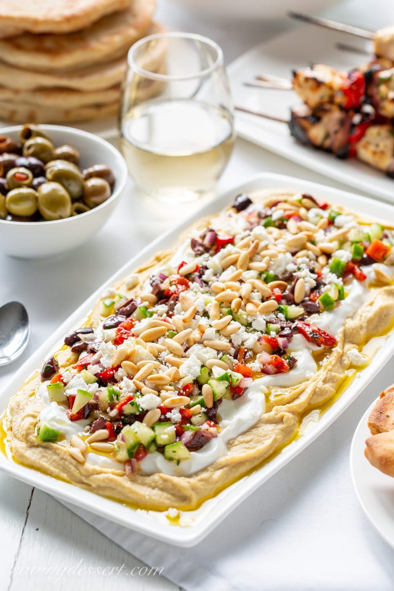 A platter of Greek Layered Hummus Dip with yogurt, olives, red peppers, pine nuts and cucumbers