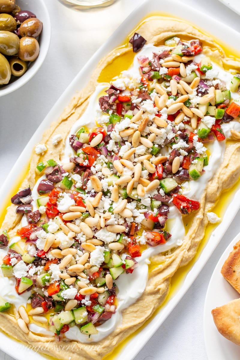 A small platter with a Greek Layered Hummus Dip with yogurt, red peppers, cucumbers, onion, olives, feta cheese and pine nuts