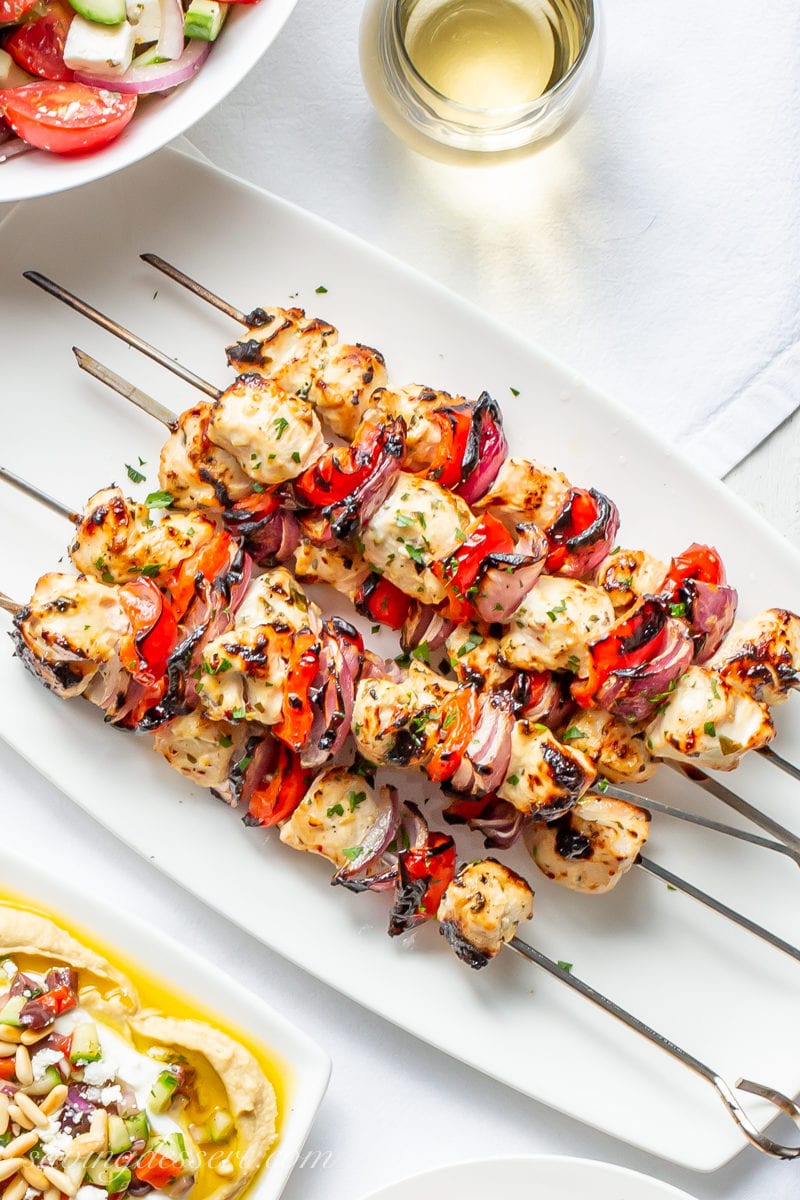 A plate of grilled Greek Chicken skewers with red peppers and purple onions served with hummus and Greek salad
