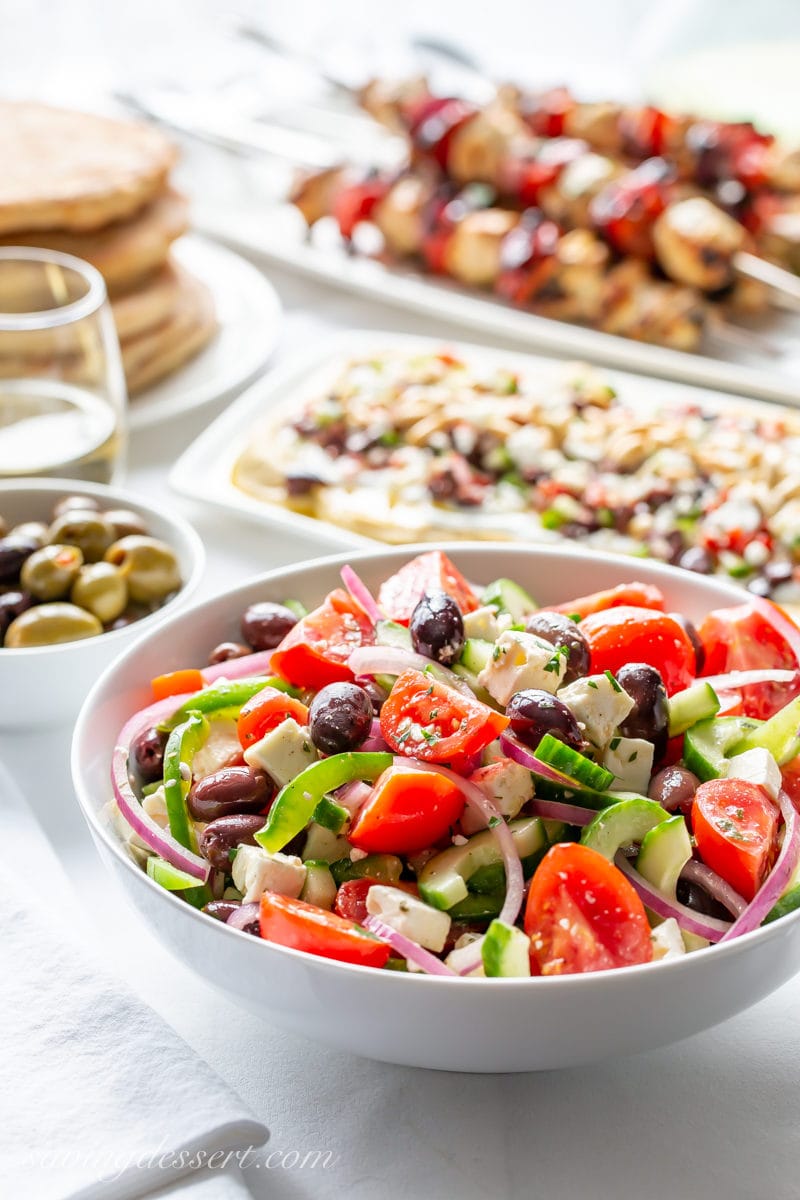 A table set with a big bowl of Greek Salad, olives, pita bread, layered Greek dip and chicken kabobs