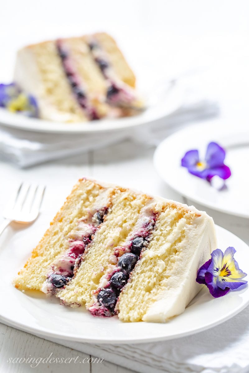 A slice of a three layer lemon cake with blueberry filling garnished with blue and yellow pansies