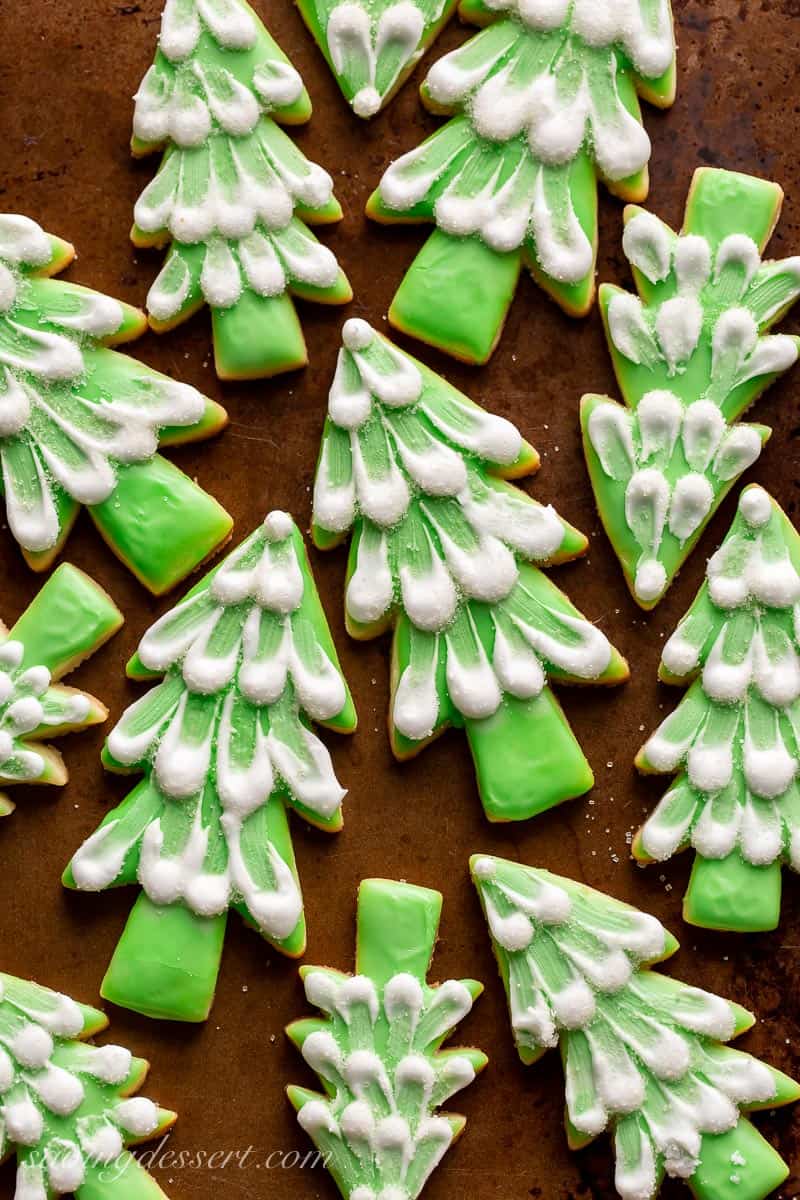 A baking sheet covered with cut out sugar cookies shaped like pine trees with green icing and white decorations