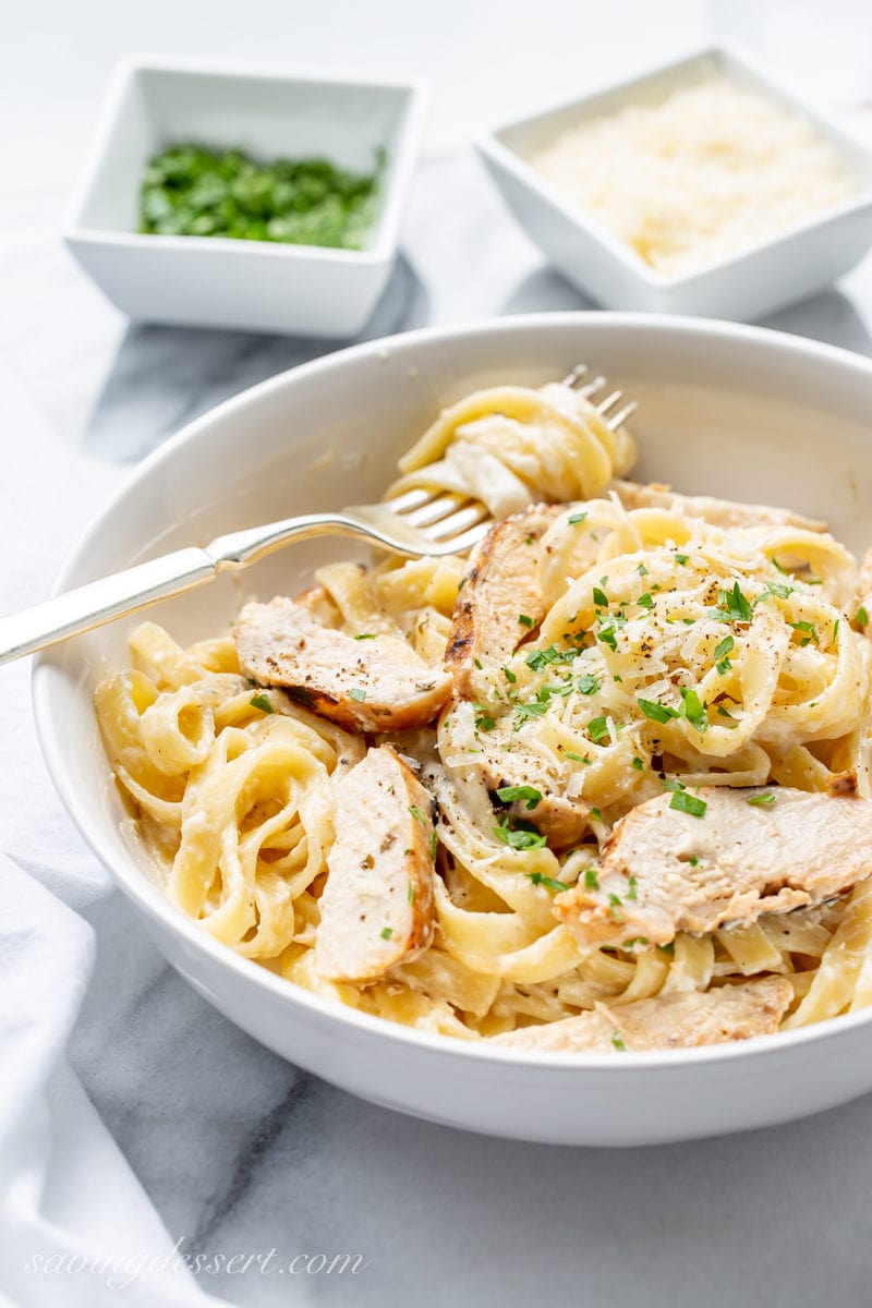 A bowl of chicken fettuccine Alfredo with bowls of chopped parsley and grated Parmesan cheese