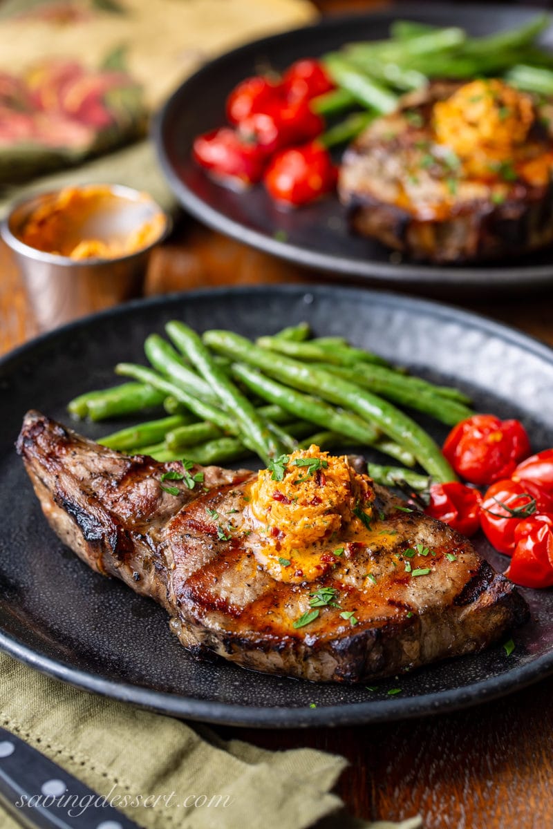 A plate with a grilled pork chop topped with chipotle butter and served with green beans and roasted tomatoes