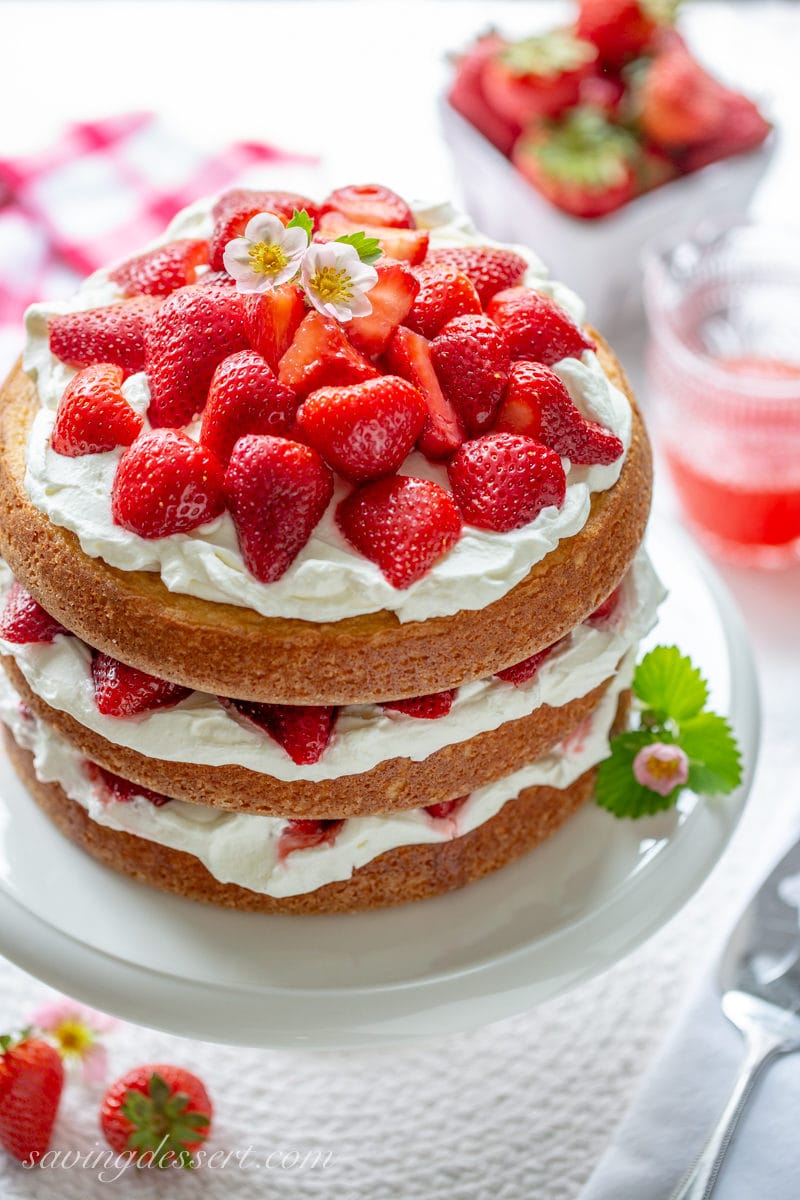 A three layer Strawberry Shortcake Cake filled with whipped cream and topped with red ripe strawberries and a few strawberry blossoms