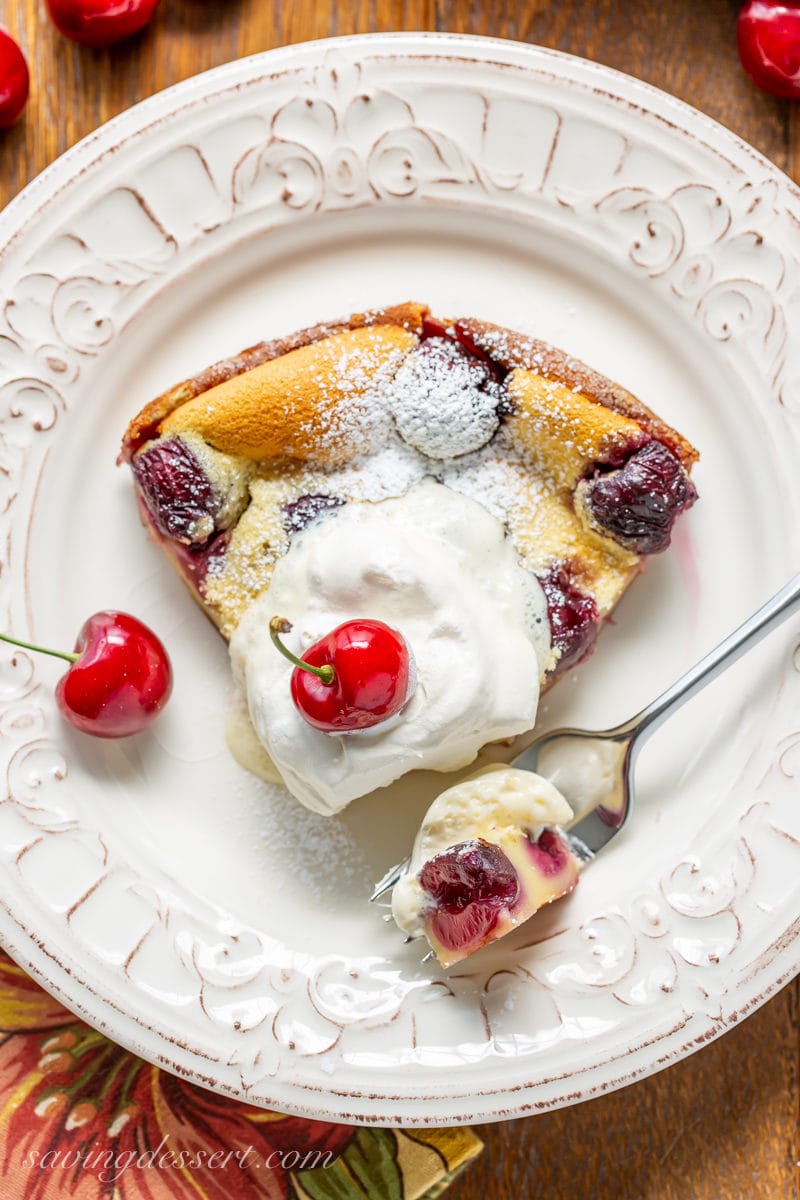 A wedge of cherry clafoutis topped with a dusting of powdered sugar and a spoonful of lightly sweetened whipped cream.