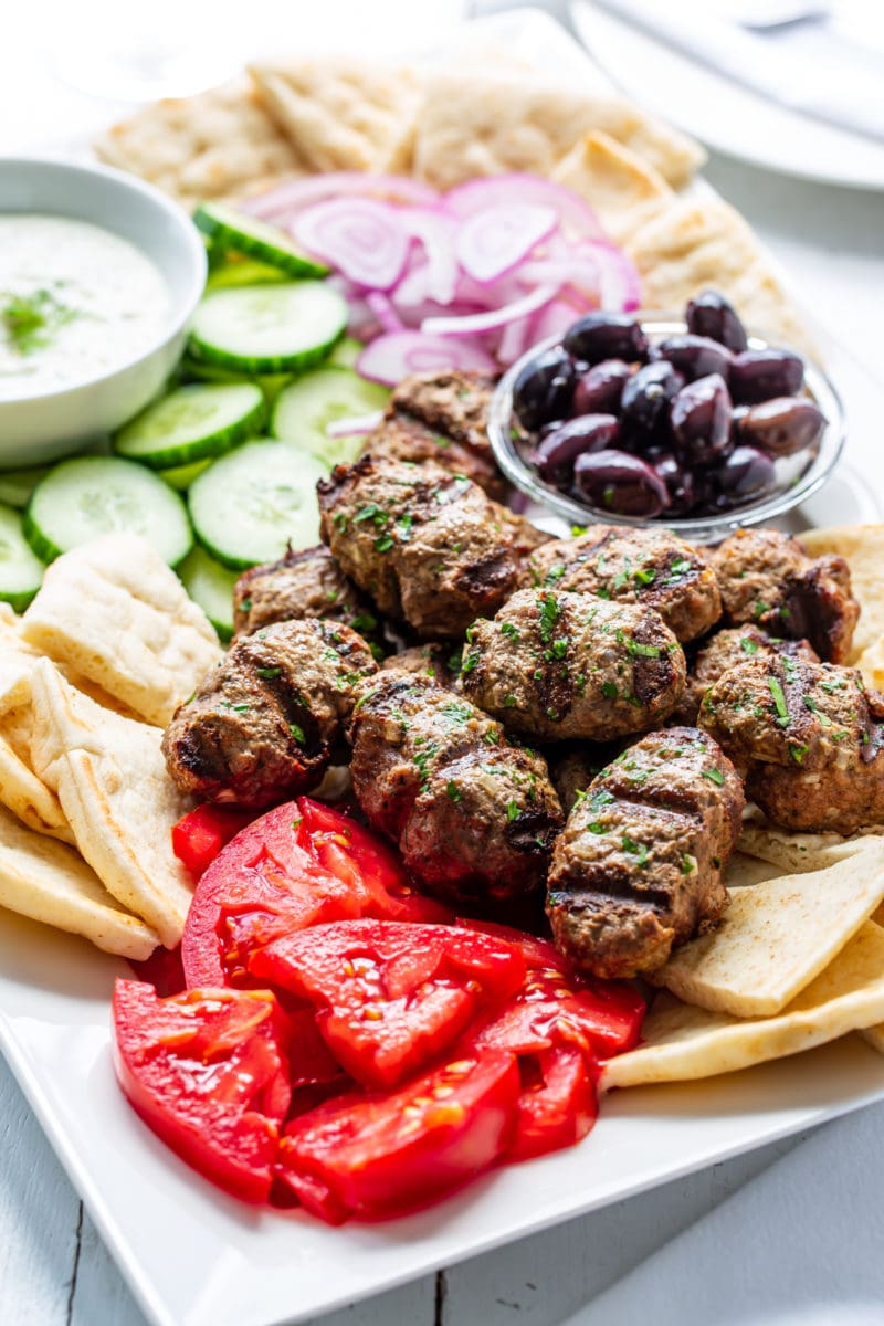 Grilled Kebabs on a platter with olives, tomatoes, pita, cucumbers and onions