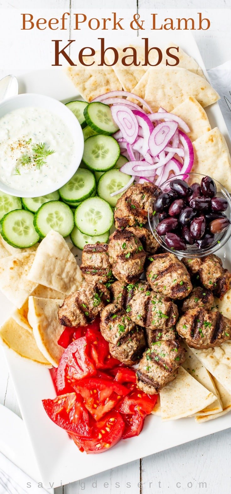 A tray with grilled lamb kebabs, tomatoes, cucumber, onion, pita wedges and cucumber-yogurt sauce