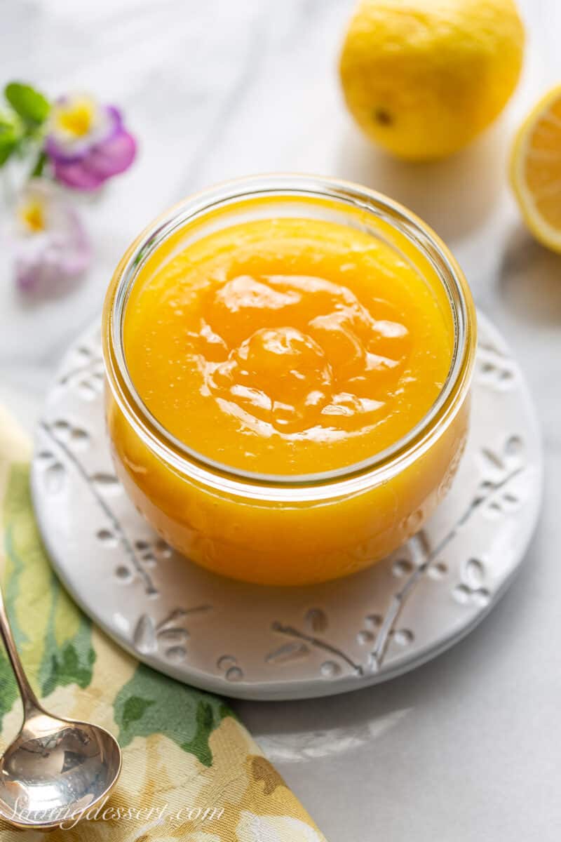 Overhead of a jar of homemade lemon curd on a plate with lemons in the background.