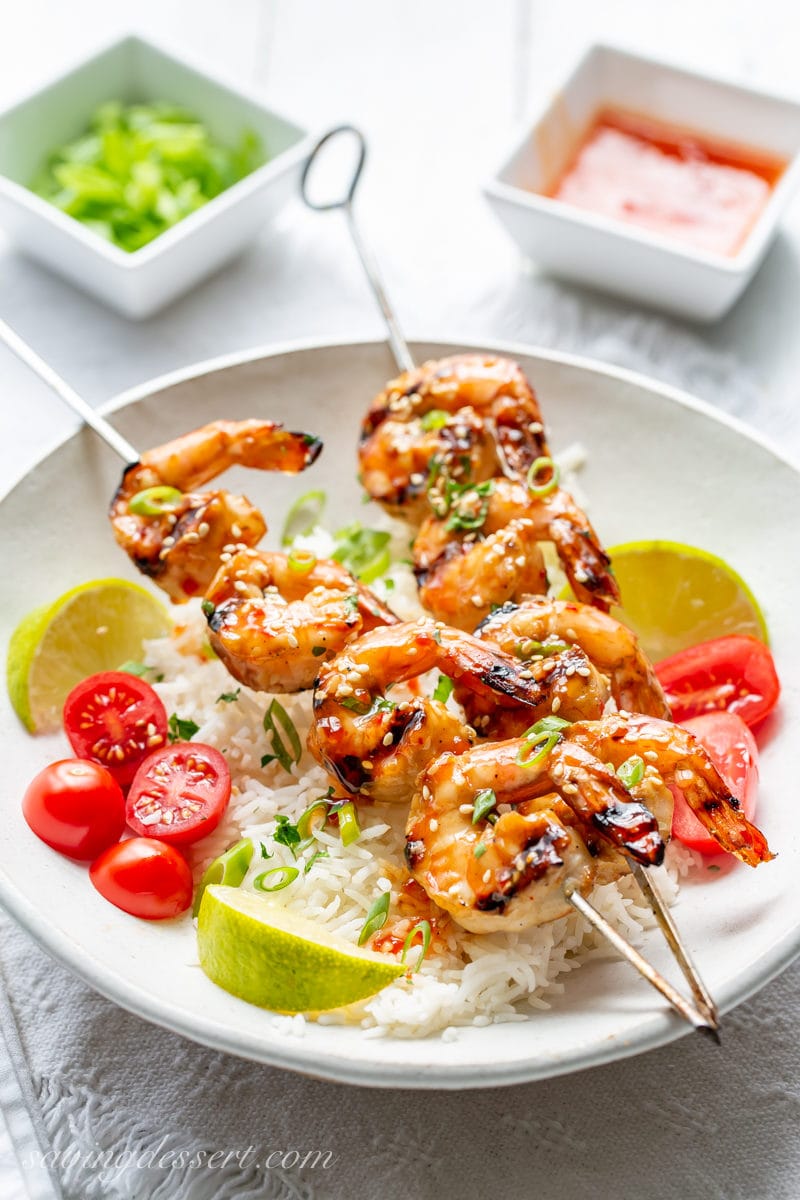 A bowl of rice with lime wedges and sliced tomatoes topped with skewers of Honey Sesame Grilled Shrimp garnished with sliced green onions.