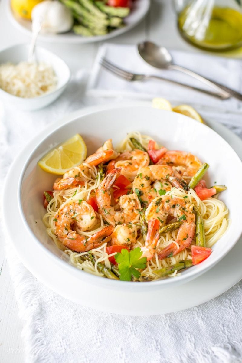 A bowl of shrimp scampi with asparagus and tomatoes over a bowl of angel hair pasta garnished with parsley
