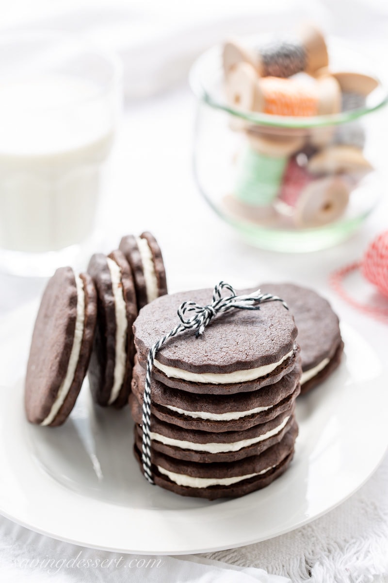 A plate stacked with homemade chocolate sugar cookies with a vanilla cream filling served with a glass of milk