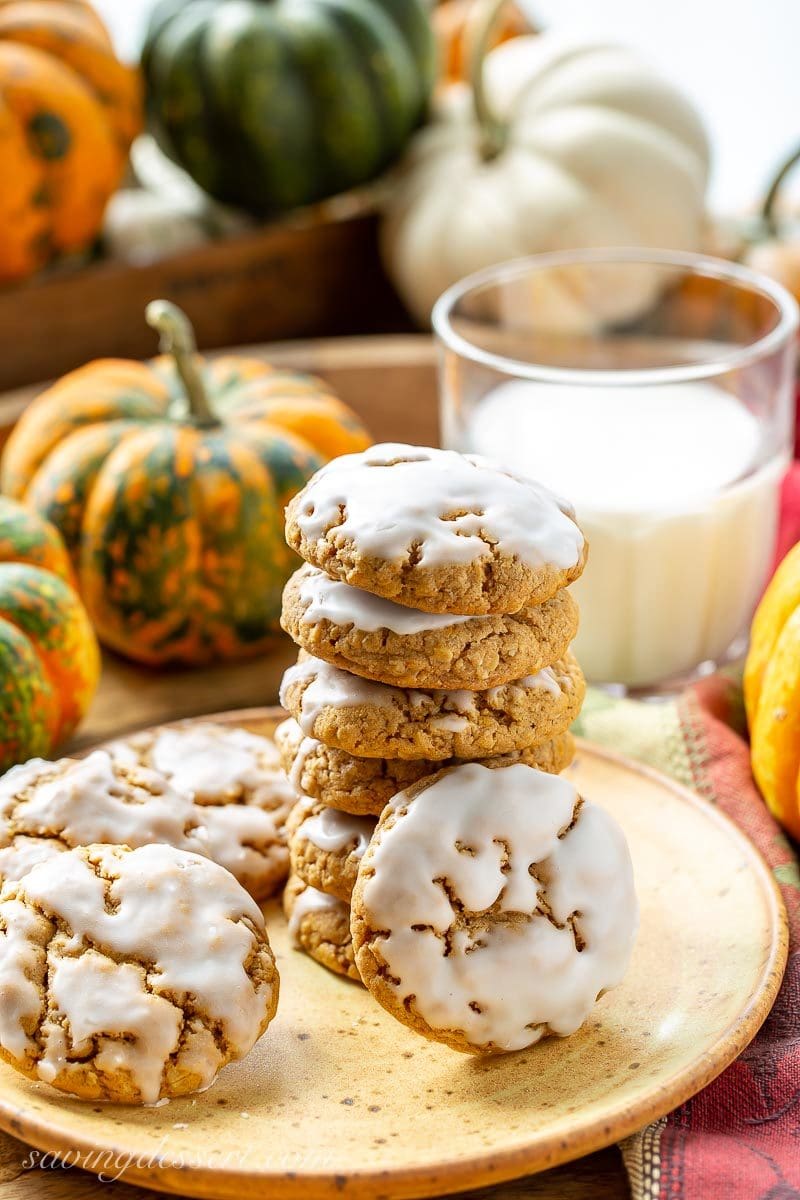 A stack of iced Spiced Pumpkin Oatmeal Cookies with a glass of milk and ornamental pumpkins