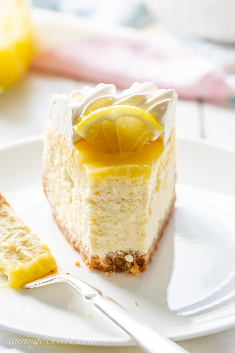 A slice of cheesecake topped with lemon curd and a sliced lemon with whipped cream