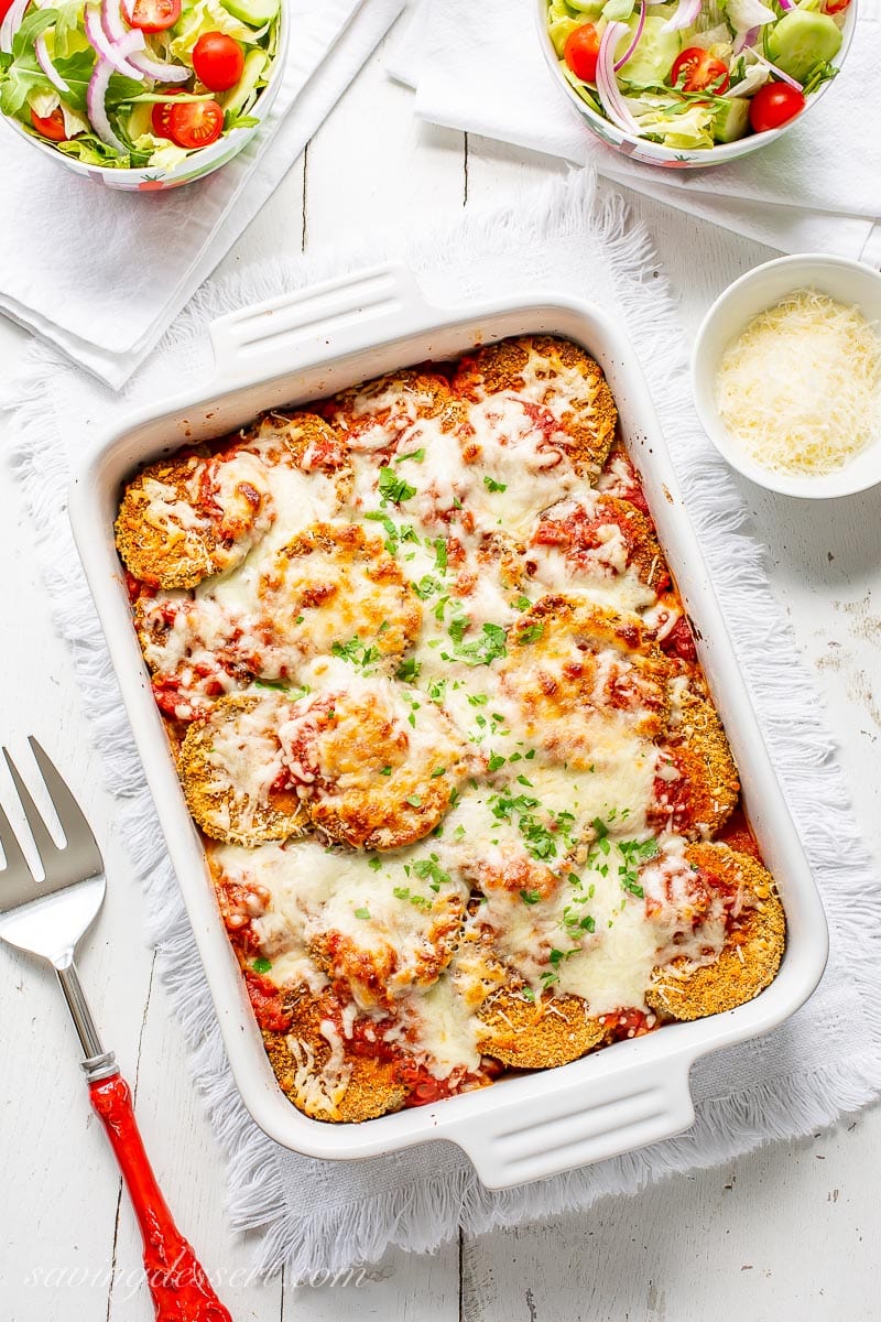 A casserole dish with eggplant Parmesan topped with mozzarella and chopped parsley. Served with extra Parmesan on the side and salad