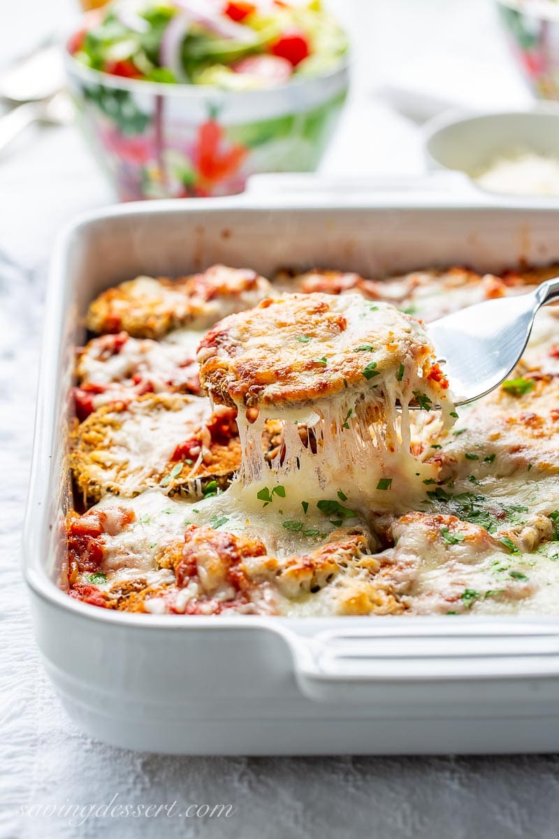 A casserole dish with a scoop of eggplant parmesan on a fork with dripping, gooey cheese