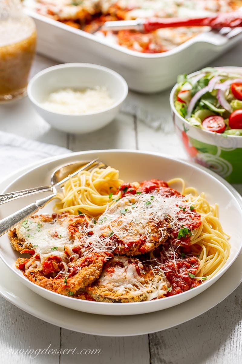 A bowl of spaghetti topped with eggplant Parmesan and served with a side salad and extra grated Parmesan in a bowl