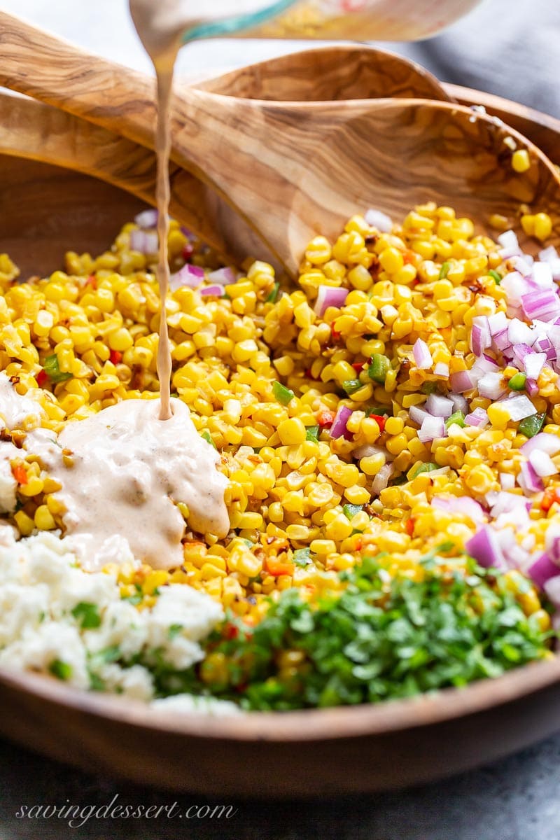 A wooden bowl of corn and onions being drizzled with a creamy dressing