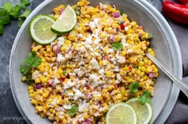 A bowl of corn salad garnished with cilantro, and fresh lime wedges