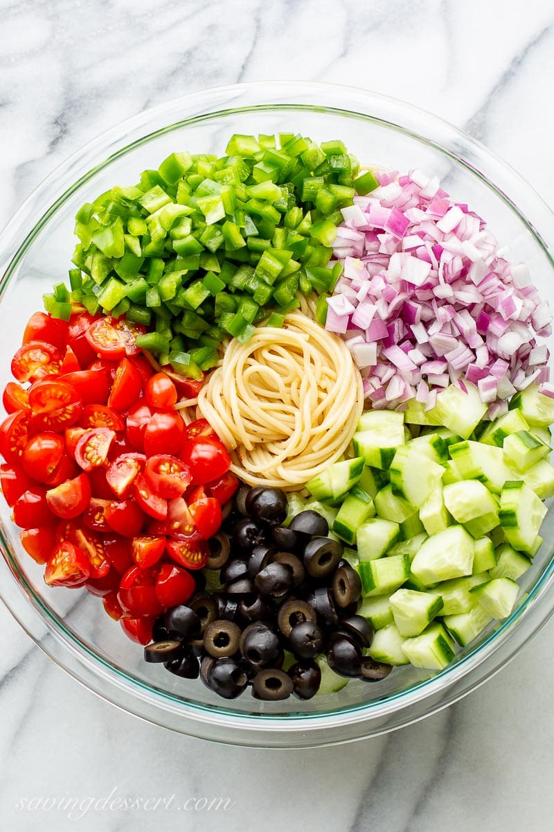 A large bowl filled with diced green bell pepper, red onion, cucumber, olives, tomatoes and spaghetti