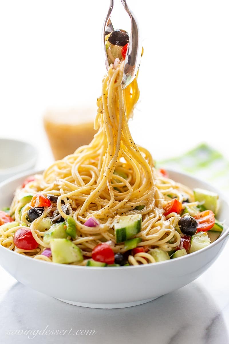 A bowl of cold spaghetti with chopped vegetables being picked up by tongs. 