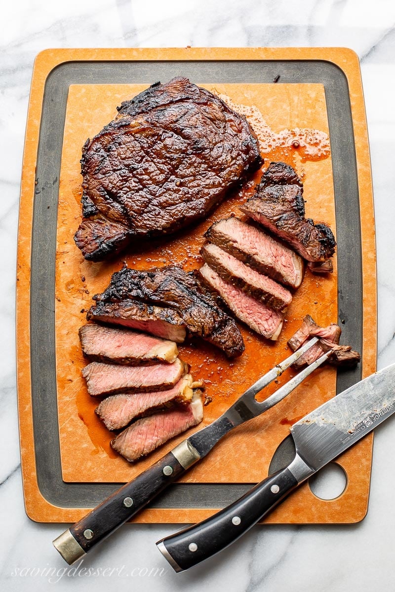 A cutting board with two grilled ribeye steaks, one sliced 