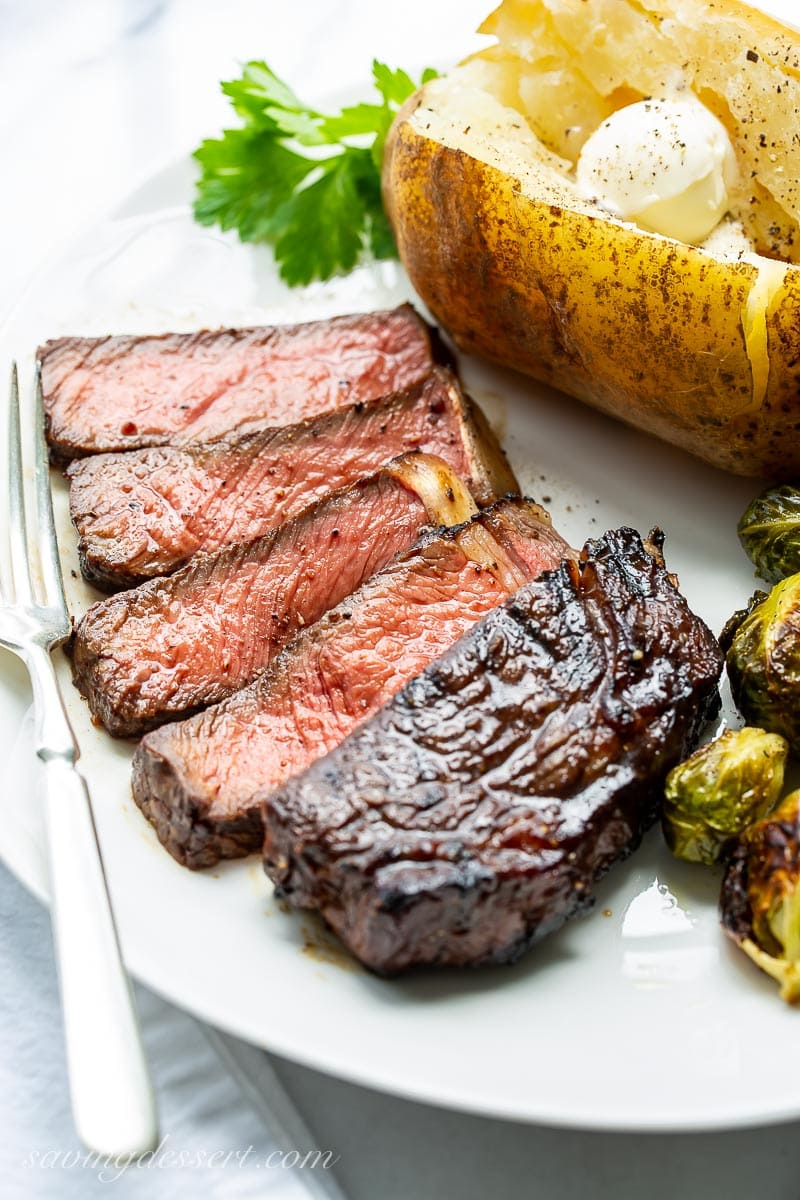 A dinner plate with a sliced ribeye steak with a baked potato and roasted Brussels sprouts. 